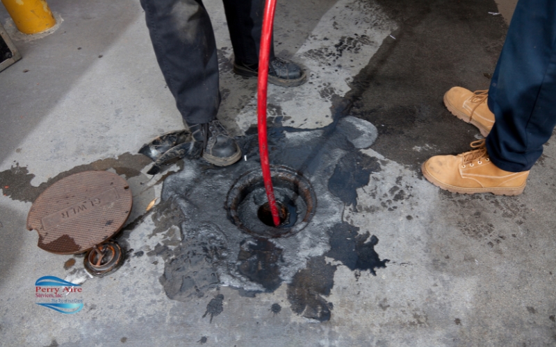 Perry Aire Services, Inc. Fredericksburg Two people standing near an open manhole with a red hose inserted, surrounded by oil stains on the ground.