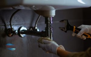 Perry Aire Services, Inc. Fredericksburg A person wearing gloves uses a wrench to fix a leaking pipe under a sink, holding a cloth to catch dripping water.