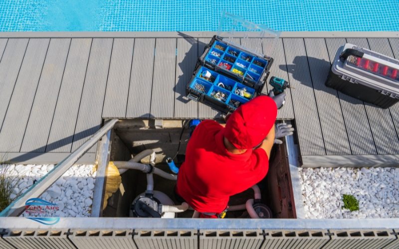 Perry Aire Services, Inc. Fredericksburg A technician in red uniform works on pool maintenance, kneeling near an open equipment hatch with tools and a blue pool in the background.